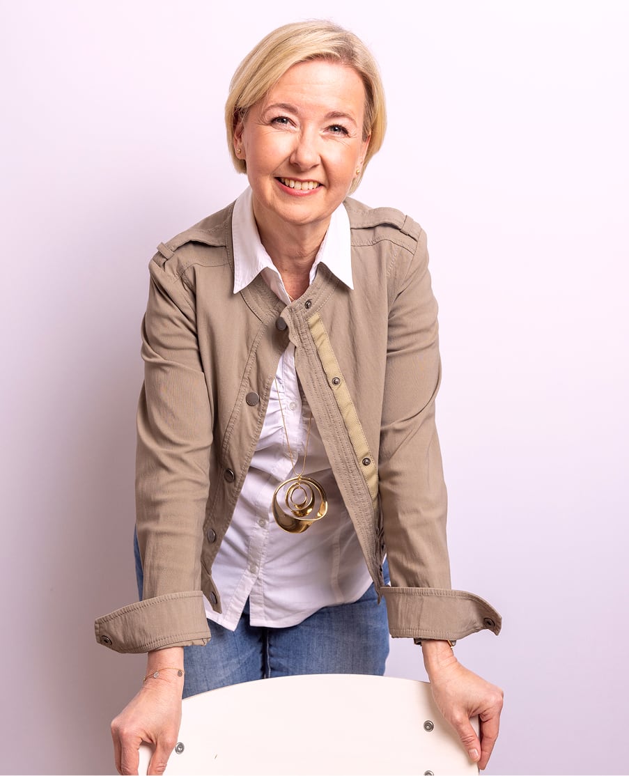 Angela Schuh-Haunold in beiger Jacke und weißer Bluse, lächelnd auf einen Stuhl gestützt