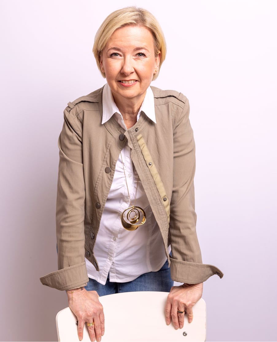 Angela Schuh-Haunold in beiger Jacke und wei&szlig;er Bluse, l&auml;chelnd auf einen Stuhl gest&uuml;tzt